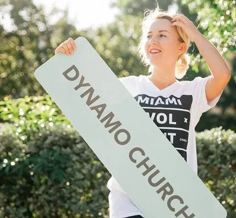 Улыбающаяся девушка держит табличку Dynamo Church на улице.