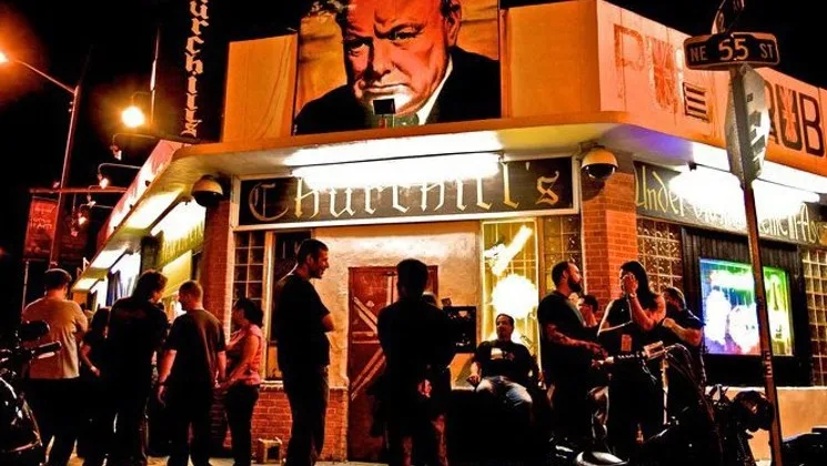 Вечерний вид на вход в паб Churchill's со стоящими снаружи людьми.
