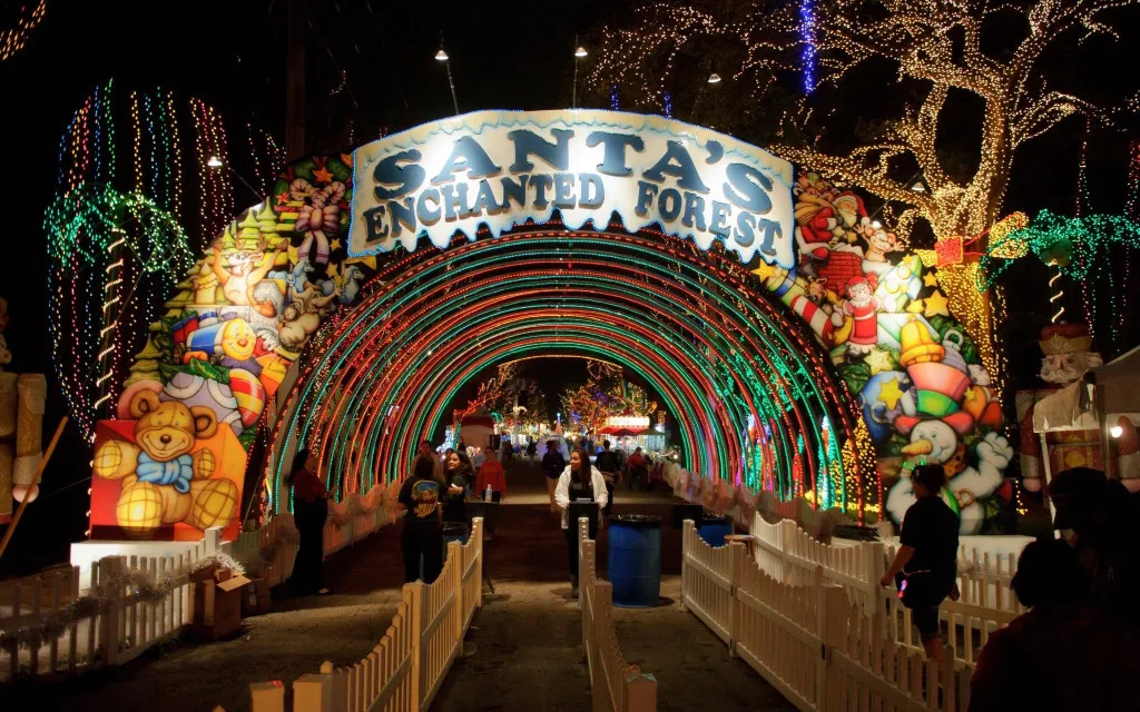 Светящаяся арка с надписью Santa's Enchanted Forest в ночном парке.