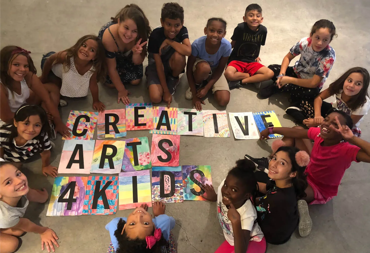 Дети сидят на полу вокруг самодельной надписи Creative Arts 4 Kids.
