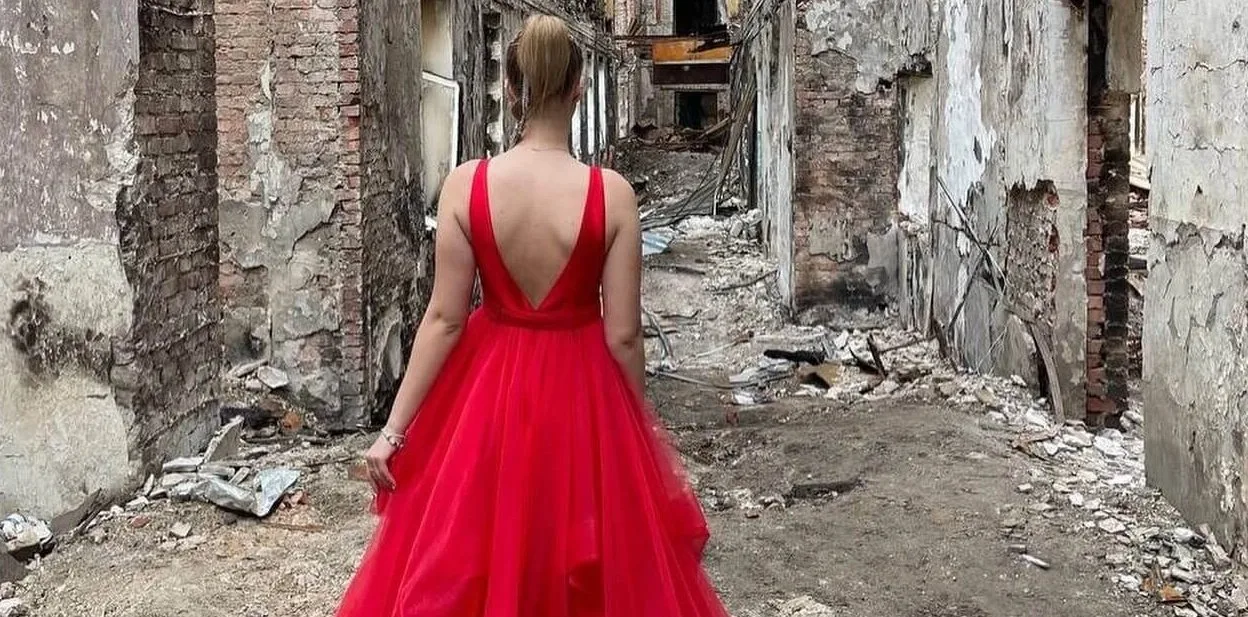 Девушка в красном платье со спины стоит среди разрушенного здания.