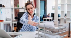 istockphoto-1130569616-612x612-e1660140442640-300x162 Young lady at desk doing paperwork and talking on smartphone.