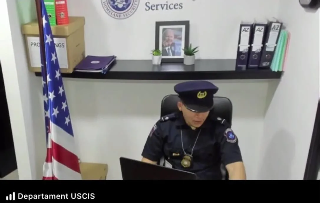 Мужчина в синей полицейской форме сидит за столом с ноутбуком под логотипом USCIS.